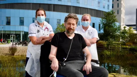 Royal Papworth Hospital Daniel Evans-Smith sitting in a wheelchair outside Royal Papworth hospital. He is looking at the camera and has two female staff members standing behind him in white uniform. They are wearing face masks. Mr Evans-Smith has an oxygen tube going into his nose and taped to the side of his face. He is wearing a black t shirt and grey jogging bottoms.