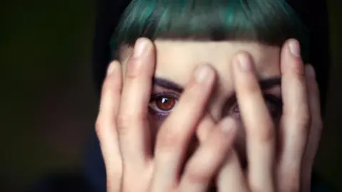 Getty Images A woman with brown eyes and dyed green hair peeks through her fingers