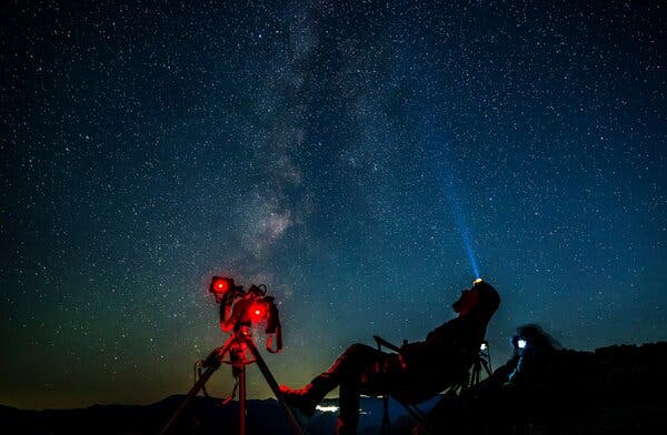 A person with a head lamp and fancy telescope/camera sits in a camping chair and looks up at the night sky with the Milky Way visible.