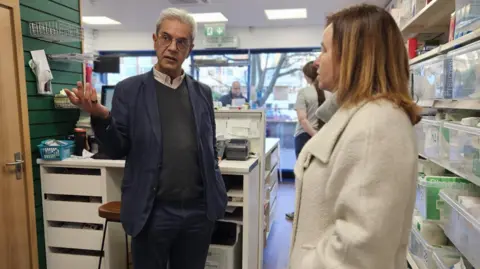 Marie Goldman Marie Goldman and Dipak Pau inside a pharmacy in Essex. Mr Pau is pointing at something while talking to Ms Goldman.