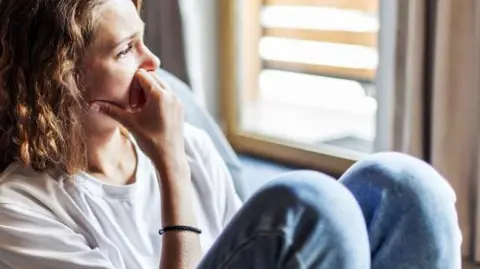 Getty Images Woman looking pensive sitting on the floor with her back against a bed