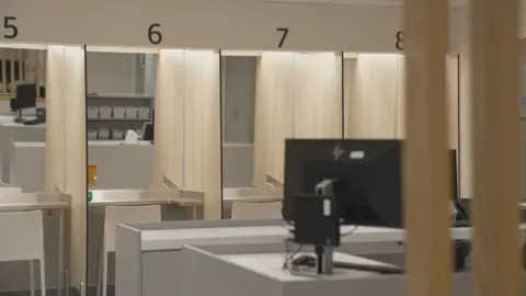 The injection booths with mirrored walls and plastic white chairs are behind a reception desk with a computer.