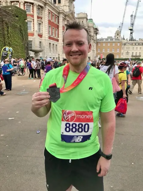 Peter Rossiter standing on the street after running a race, wearing a lime green sports top and black shorts. He is holding up the medal which is on a lanyard around his neck.