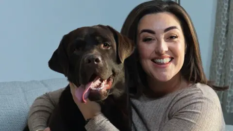 Phil Tragen/The Christie/PA Media Bianca Perea with long brown hair smiling wearing a beige long sleeved t-shirt cuddling her brown Labrador dog as they sit on a cream sofa