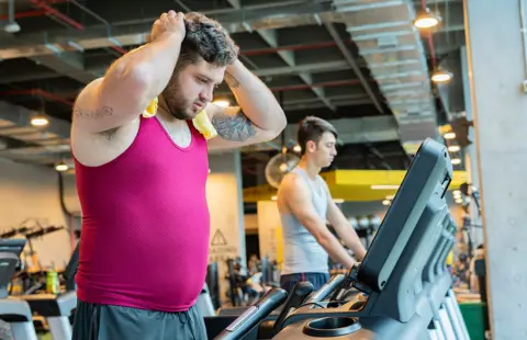 Getty Images Two men on exercise machines in the gym, the man in foreground is using a flannel to wipe up sweat from working out, with his hands on his head.