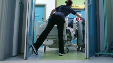 Getty Images A member of hospital staff holds open a door with her foot while wheeling a patient on a trolley