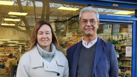 Marie Goldman Marie Goldman and Dipak Pau wearing coats outside a pharmacy in Essex. They are both smiling at the camera