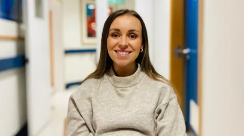 Steph Dennison is sitting in the hospital ward. Wearing a soft grey jumper, she is smiling.