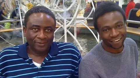 Femi Akinnola and his son Lobby are pictured sitting by a fountain, looking into the camera. Femi is wearing a blue t-shirt while Lobby is sitting next to him, wearing a light grey sweatshirt.