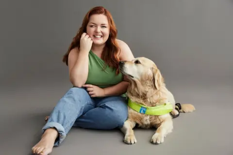 RNIB Lucy Edwards is sitting on the floor with her guide dog, Molly, to mark her becoming an RNIB ambassador. She has long red hair and is smiling. She is wearing a sleeveless green top and jeans 