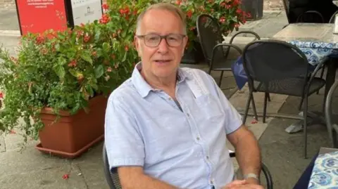 University of Aberdeen Man, Brian Watt, smiling at looking at camera, wearing a short-sleeved light blue shirt, he appears to be sitting in an outdoor cafe.
