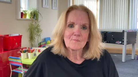 A woman sitting looking at the camera with a neutral expression. She is wearing a black top. She is in a white room with some pictures on the wall. There are some plants on a windowsill while the blinds of a far window are closed. Behind her is a child's chair and table with some pots of pencils on it. Also in the background is a table football