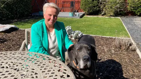 A woman sitting outside on a metal chair, next to a metal table. She has short blonde hair and is wearing a white top and green jacket. She is smiling at the camera. Next to her is a large black dog. Behind her is an area of woodchip and behind that an area of grass. In the background a wooden fence and some hedge.