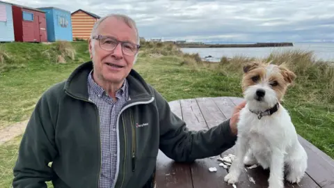 BBC Brian Watt, smiling man with small dog which is sitting next to him, with the sea and beach huts in the background