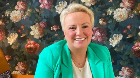 BBC A woman with short blonde hair wearing a green jacket and white top smiling at the camera. She is sitting on an orange armchair in front of wall paper with a flower pattern.