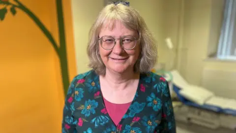 Professor Zosia Miedzybrodzka, woman smiling and looking at camera, wearing a bright floral top, with a hospital bed behind her.