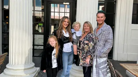 Chanel Williams James Lee Williams poses with his mum, nieces and sisters outside a restaurant doorway. The family are smiling and looking at the camera. 