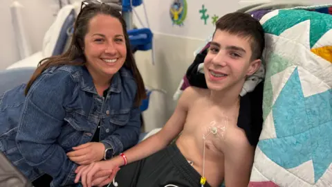 Steve Hubbard/BBC Finley and his mum Rhys in a hospital room in Broomfield Hospital. Finley is lying upright on the hospital bed, he is not wearing a top as he has a medical tube running into his chest. He is lying on top of a multi-coloured patchwork blanket, and he wears black tracksuit bottoms. He is holding hands with his mum Rhys who is sat beside him. Rhys has a denim blue jacket on and has dark brown hair running down past her shoulders. She has a pair of sunglasses sitting on top of her head, and both are smiling at the camera