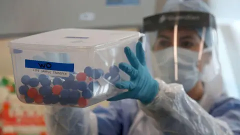 Getty Images An NHS worker is holding a box of coronavirus tests. She is wearing a visor, gloves and an apron. You cannot see her face because she is also wearing a mask.