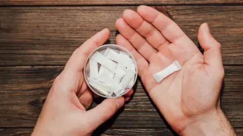 Getty Images A white circular tub containing nicotine pouches being held in one hand, with a single pouch in the other hand. The pouch is a small rectangle which fits easily in the palm of a hand.