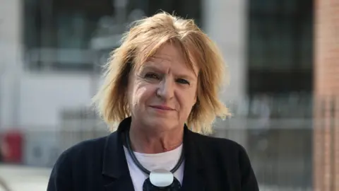 Jamie Niblock/BBC Head and shoulders photo of Deborah Coles. She has straight blonde shoulder-length hair with a centre parting. She is wearing a dark jacket with a white top underneath, and a large black and silver necklace. She is standing in front of a building with railings outside, and is looking directly into the camera