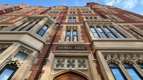 Stuart Woodward/BBC Arundel House in London. The building is a mixture of red bricks and sandstone window frames and door frames. The words Arundel House are written on a dark sign over the top of a closed wooden door. The photo is taken from a low angle looking up at the building, with blue skies above.
