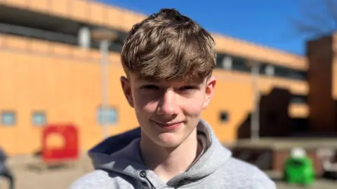 Lee looks directly at the camera with a blurred-out building in the background. He has short brown hair and is wearing a grey hoodie.