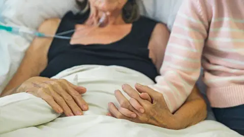 Getty Images An elderly woman lays on a hospital bed. You can't see her face but one of her hands is being held by someone younger wearing a pink and white striped jumper.