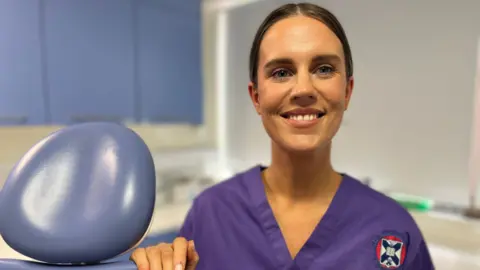 Zoe standing in a dentists' room, with her right hand resting on a blue dentist's chair. She is wearing purple scrubs and has brown hair which is tied back.