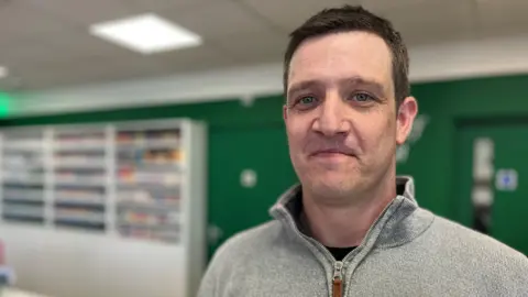 Alan McLevy looks directly at the camera, standing in front of a dark green wall. He is wearing a grey fleece and has short brown hair.