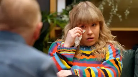 BBC / Twenty Twenty Productions Ltd Nicole in therapy, long blonde hair, wearing a striped knitted top, against a blurred interior background