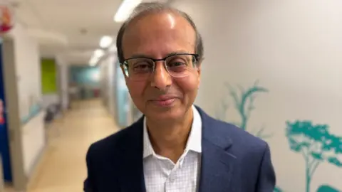 Steve Hubbard/BBC Dr Manas Datta wears a white shirt with light checked pattern, and a navy blue suit jacket over the top. He is stood in a hospital corridor with white walls behind him and turqouise-coloured stencils of plants on the walls. Dr Datta has dark thinning hair and wears spectacles. He is smiling at the camera.