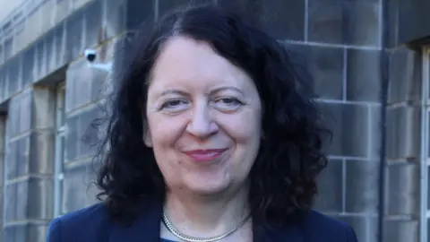Ash Scotland Sheila Duffy is standing in front of a grey sandstone building, looking directly at the camera. She has shoulder length curly black hair and is wearing a blue jacket.