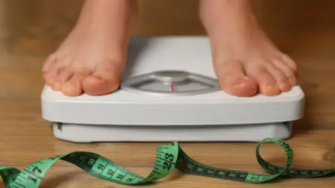 Getty Images Bare feet on a set of scales with a twirl of green tape measure in the foreground