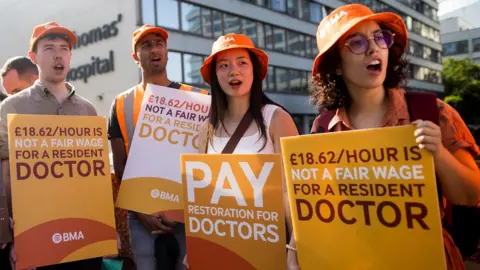 Getty Images Four doctors on strike, wearing orange BMA bucket hats and holding up placards calling for more pay, saying that '£18.62 per hour is not a fair wage for a resident doctor'. In the background St Thomas' Hospital is visible. Taken in London in July.