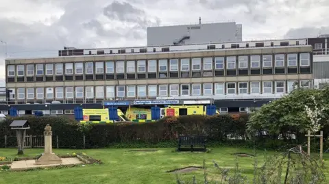 David Robinson / Geograph A long, wide building with numerous windows stands in the centre, with ambulances outside an entrance, a sign reads 'Darlington Memorial Hospital'. A park sits in front. 