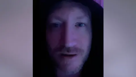 Leicestershire Police Paul Lipscombe looks straight ahead. He has a moustache and goatee beard. His face is in shadow and he appears to have a hood pulled up on his head