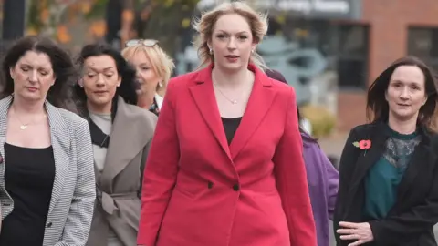PA Media A group of women arrive at the tribunal office. At the head of the group is Bethany Hutchison, who has long blond hair and is wearing a red coat. Beside her are several smartly dressed women with long brown hair.