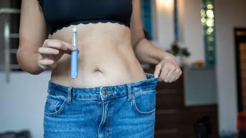 Getty Images A woman shows her weight-loss progress by holding a GLP-1 injection pen in one hand and pulling on the waistband of jeans that no longer fit