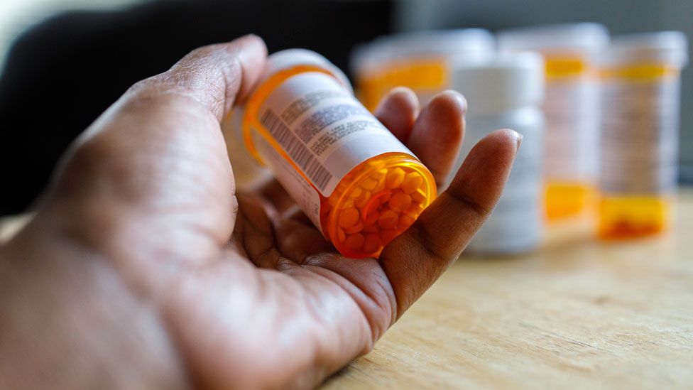 A hand holding a cylindrical bottle of pills. It's light orange in colour, with a white lid and white barcode label. We can see lots of small round pills through the bottom of the translucent container. In the background, we can see the blurry outline of several similar pill bottles.