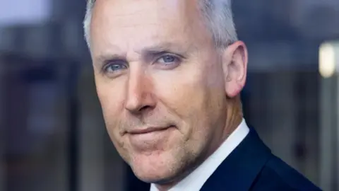 Blavatnik School of Government A facial close up of Ciaran Martin looking directly into the lens of a camera. He has got white short hair and is wearing a white shirt underneath a navy suit. The background is blurred.