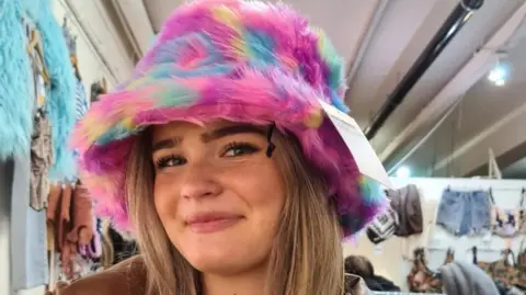 Nikki Fox/BBC A smiling Amelia Butlin standing in a clothing shop posing at the camera in a multi-coloured fur hat with the price tag attached.