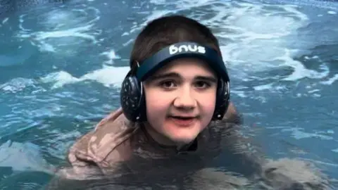 Family Tobie, in a swimming pool facing the camera. He is wearing a brown t-shirt and black ear defenders