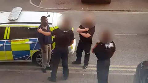 Handout A photo taken from a window above of a police officer with his hand on Valdo Calocane's shoulder, next to a police car, with two more officers standing nearby