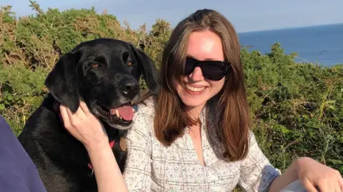 Gareth Williams Rhiannon has long dark hair and is wearing a patterned white top and sunglasses. She is sat next to the family's black labrador, Jet, near Langland in the summer of 2022.