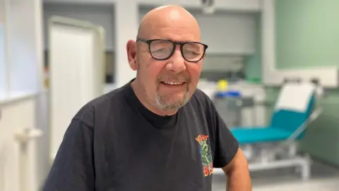 BBC Phillip Marks wears a black t-shirt and black glasses. He smiles as he perches on a trolley in a doctor's treatment room.