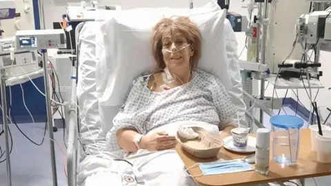 Lynda Sawyer Lynda Sawyer wearing a hospital gown and propped up by pillows in a hospital bed surrounded by medical equipment. She is wired up to monitors and has a nasal cannula for oxygen. There are various items on the wooden tray positioned over one side of the bed, including a water jug and cups.