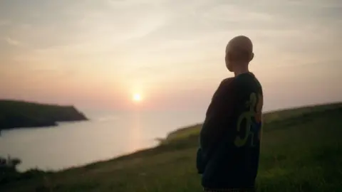 BBC/October Films/Neil Bonner Kris is standing on the coastline. She is wearing a cardigan and is looking out on to the horizon. The Sun can be seen shining in the distance.