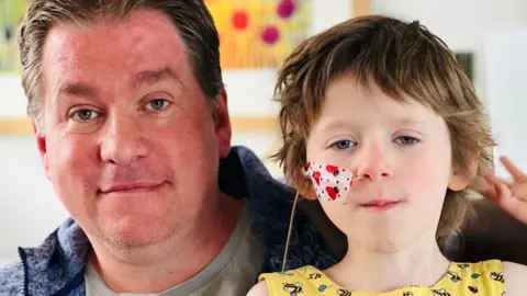 Mike Ashmore and his daughter Annabel. Mike has dark hair and Annabel has short brown hair and tube coming out of her nose held by a plaster with ladybirds on it.