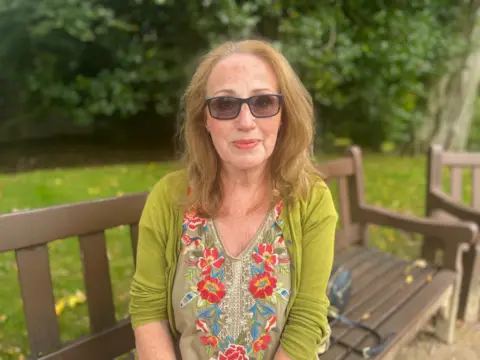 May Hooper wears a lime green cardigan and floral top, sitting on a bench in a park